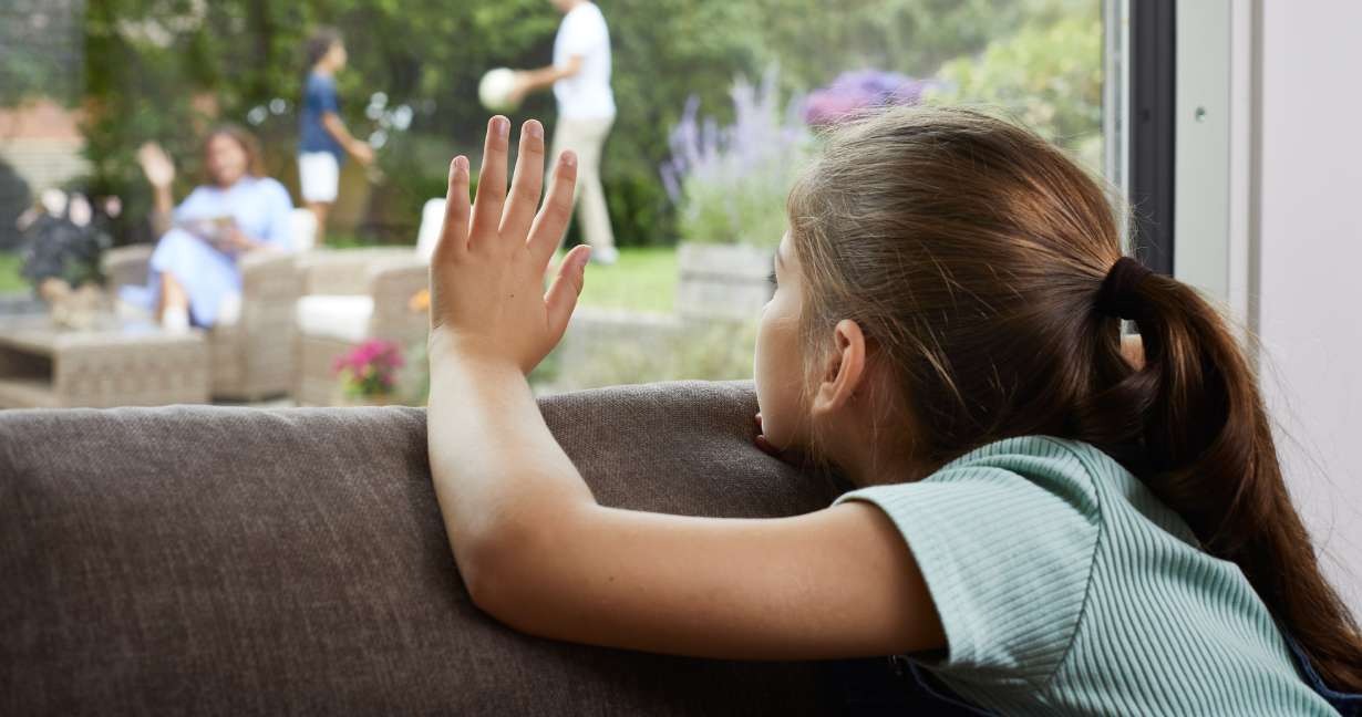 En pige sidder i en sofa og kigger ud ad vinduet, mens hun vinker til sin familie. Pollenallergi kan betyde, at børn må holde sig indenfor på dage med høje pollental.